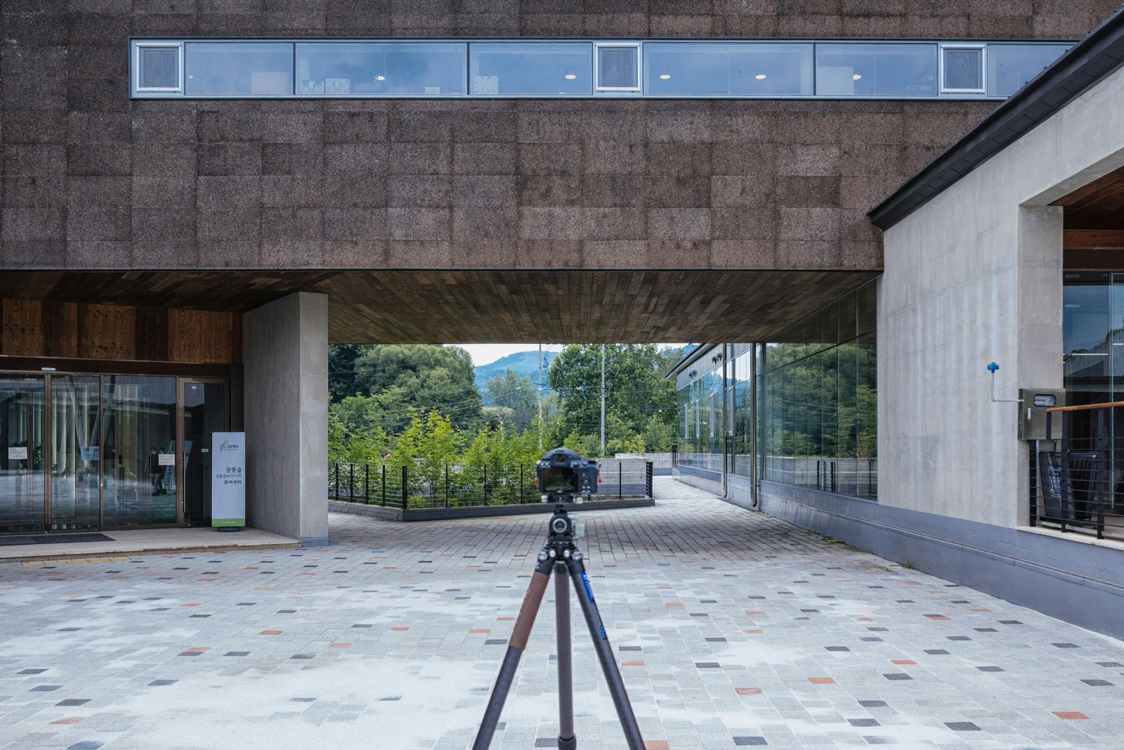 A small camera on a tripod in a paved courtyard beneath a modern concrete and wood building