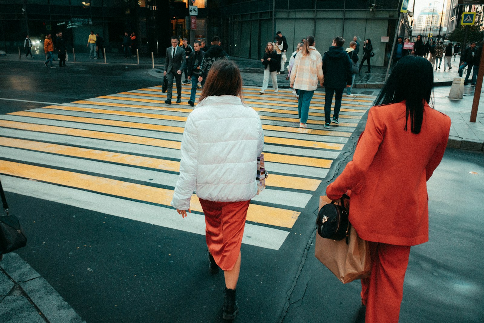 Two women in white and orange jackets crossing a yellow-striped crosswalk in a busy city, shot on a Ricoh GR